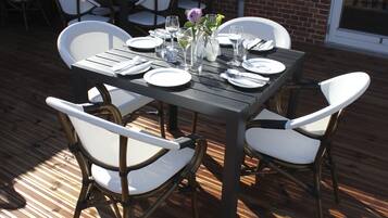 Mesa de comedor al aire libre con sillas blancas, platos, vasos y un jarrón con flores.