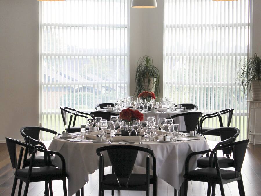 Uma sala de jantar com uma mesa redonda posta para seis pessoas, rodeada por cadeiras pretas, sob iluminação pendente, com grandes janelas e vista para o exterior.