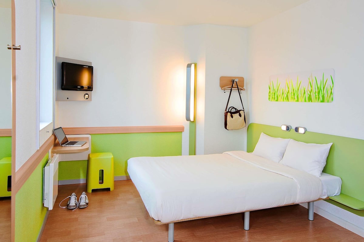 Ein Hotelzimmer mit einem Bett, einem Schreibtisch mit einem Laptop, einem Fernseher, einer Wandlampe, einer Handtasche und einem grünen Wandbild.