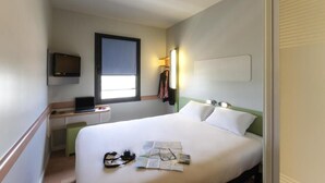 Een hotelkamer met een bed, een bureau met een laptop, een aan de muur bevestigde televisie en een raam dat natuurlijk licht biedt.