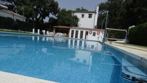 Una piscina con agua azul cristalina, rodeada por una solana con baldosas. Se ven sillones de playa y sombrillas al fondo, y un edificio con tejado y ventanas de cerámica.