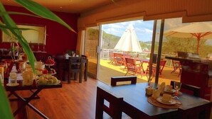 A dining area with a table set for two, a large window with a view of the outdoors, and a red wall. - Villa Moringa Guesthouse (Windhoek)