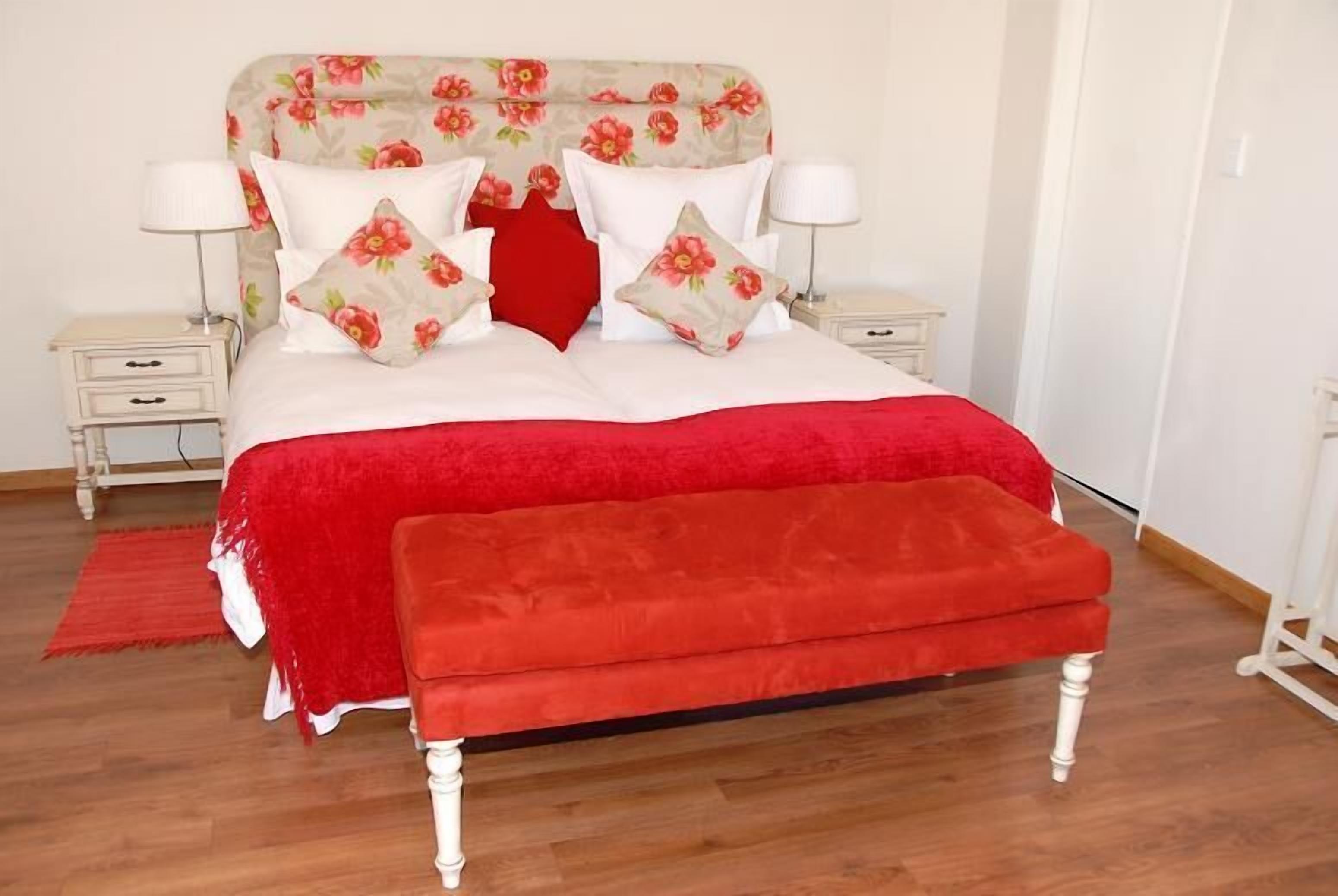 A neatly made bed with a floral headboard, white bedding, and red accents. There are two bedside tables with lamps, a red rug, and a wooden bench at the foot of the bed.