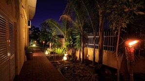 A nighttime view of a narrow pathway flanked by buildings and trees, with ambient lighting and a small garden area. - Villa Moringa Guesthouse (Windhoek)