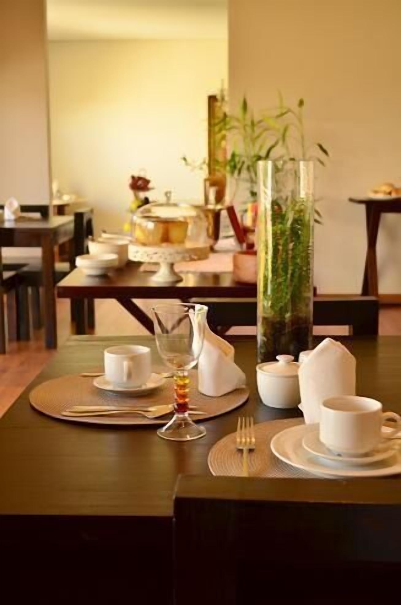 Una mesa de comedor con platos blancos, tazas y una jarrón, con un florero de plantas verdes al fondo.