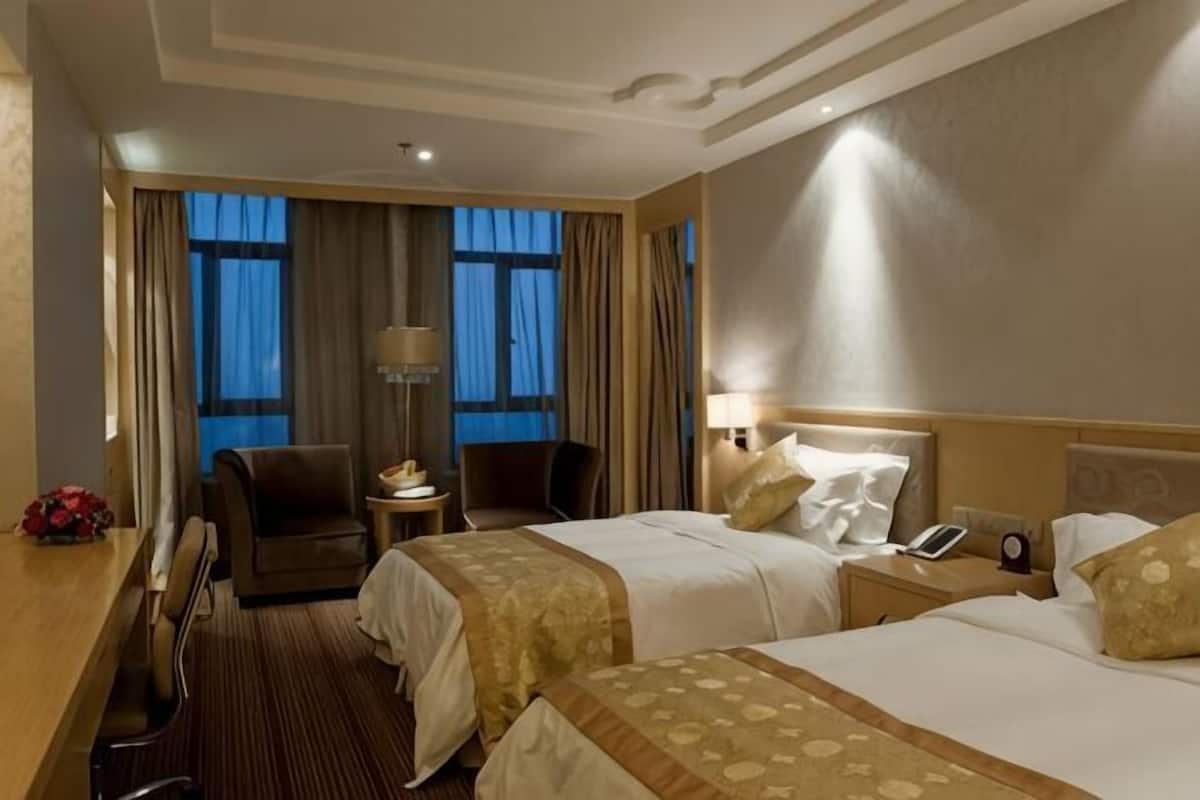 Habitación de hotel con dos camas, un escritorio, una silla, una lámpara y un ventanal con cortinas.