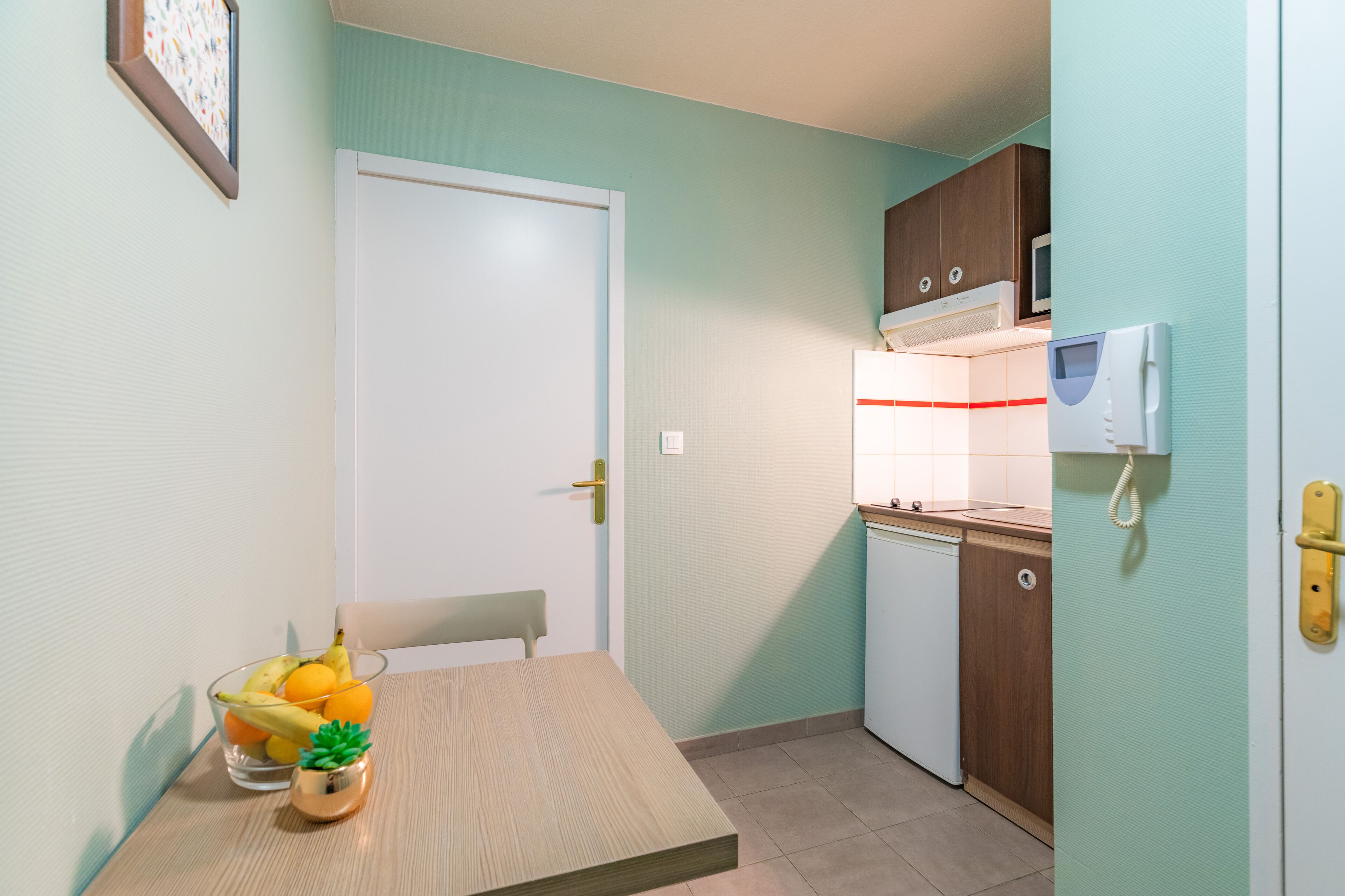 A compact kitchen with a microwave, a small refrigerator, and a sink. There is a dining table with a bowl of fruit and a small potted plant.