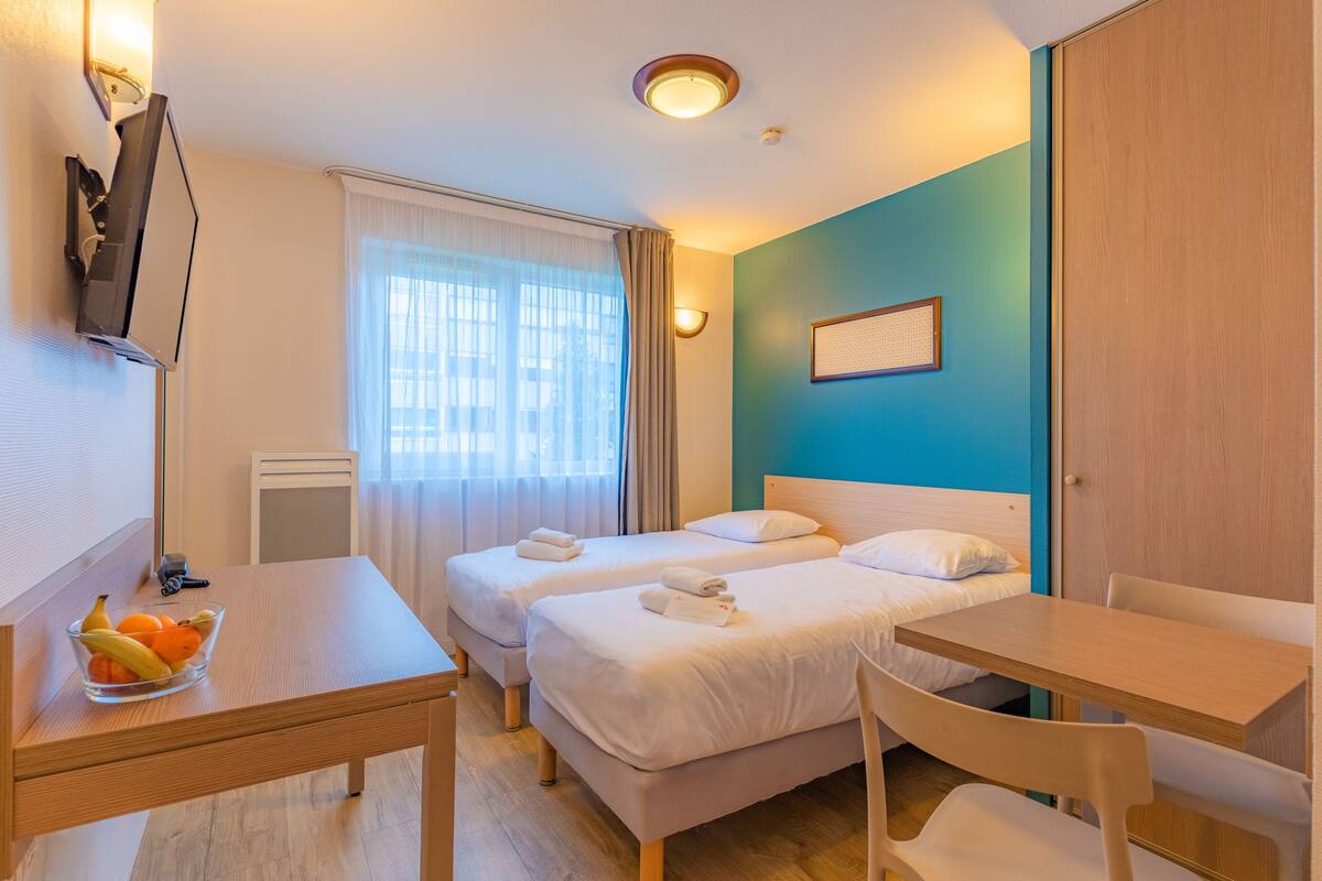 A hotel room with two beds, a wooden table, a bowl of fruit, and a TV on the wall.