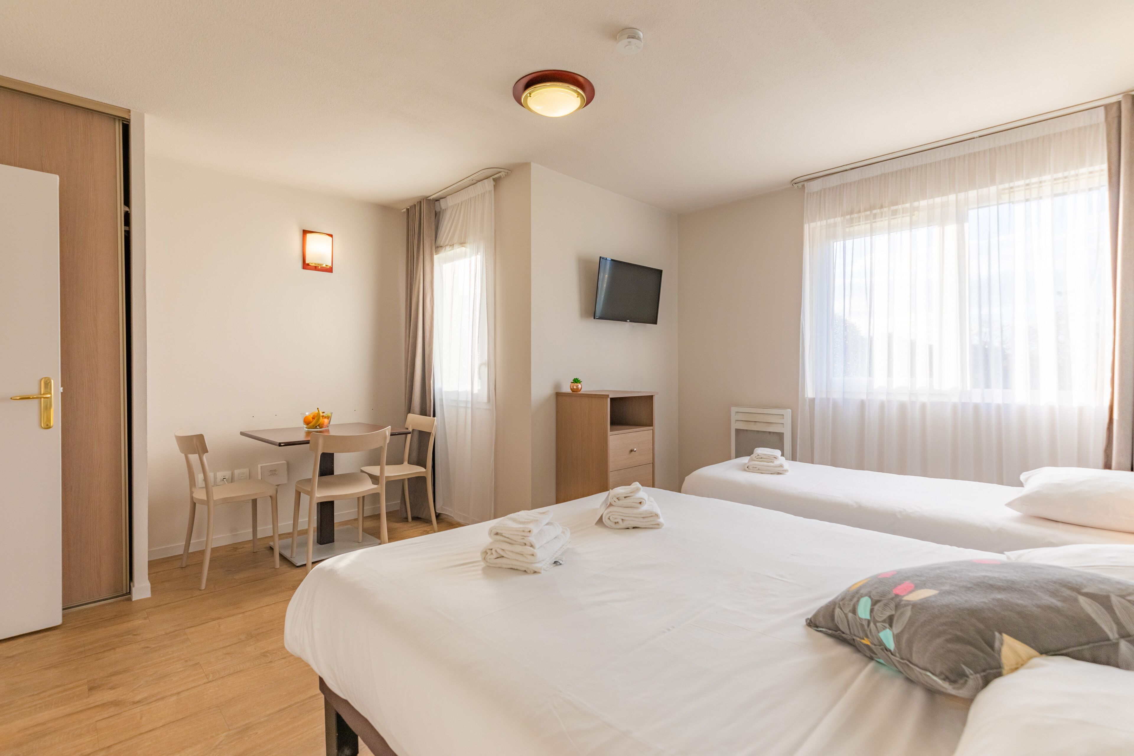 Habitación de hotel con dos camas, un escritorio con sillas, un televisor montado en la pared y una ventana con cortinas.