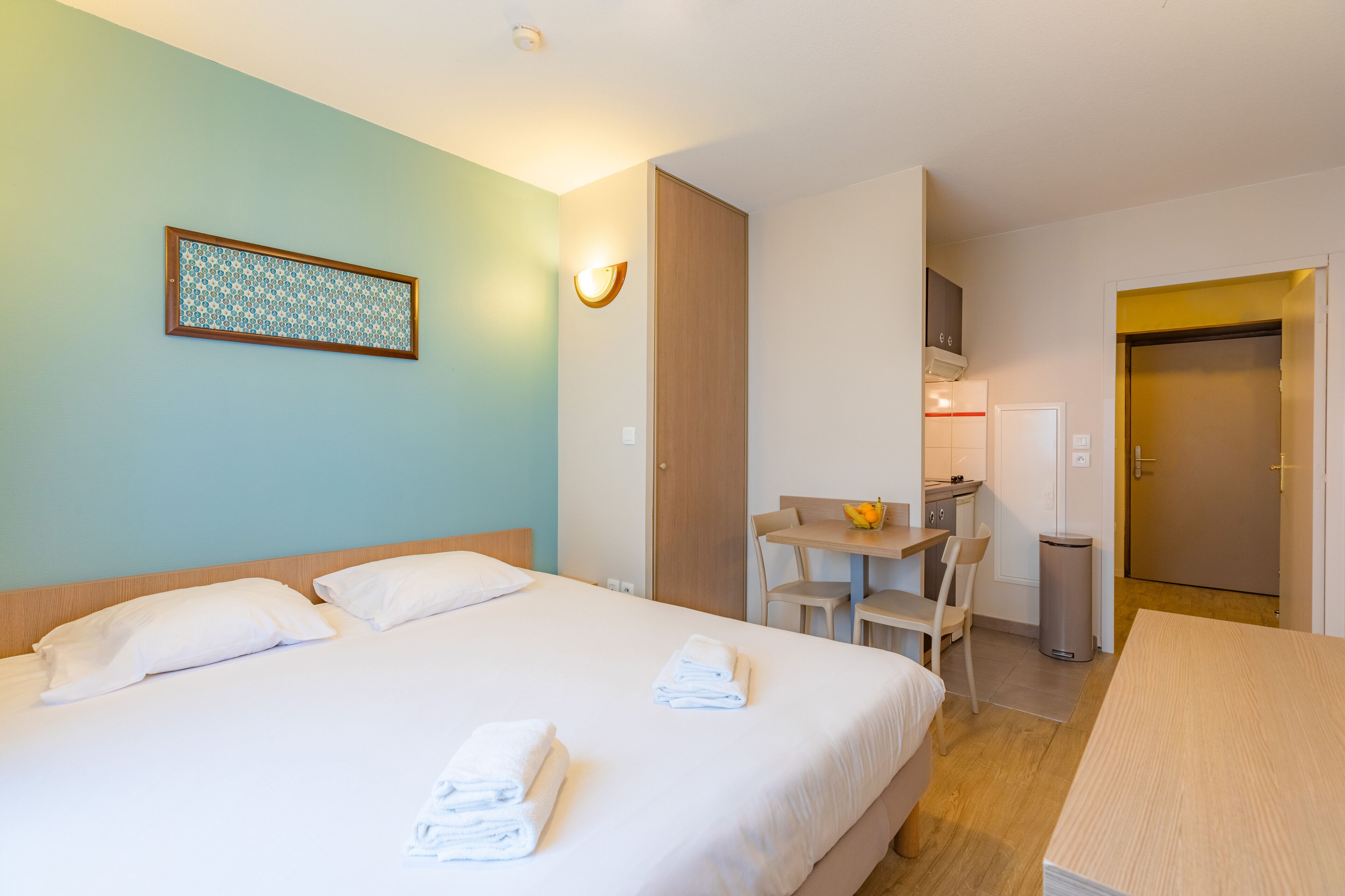 A hotel room with a bed, a desk, a chair, a small kitchenette, and a bathroom.