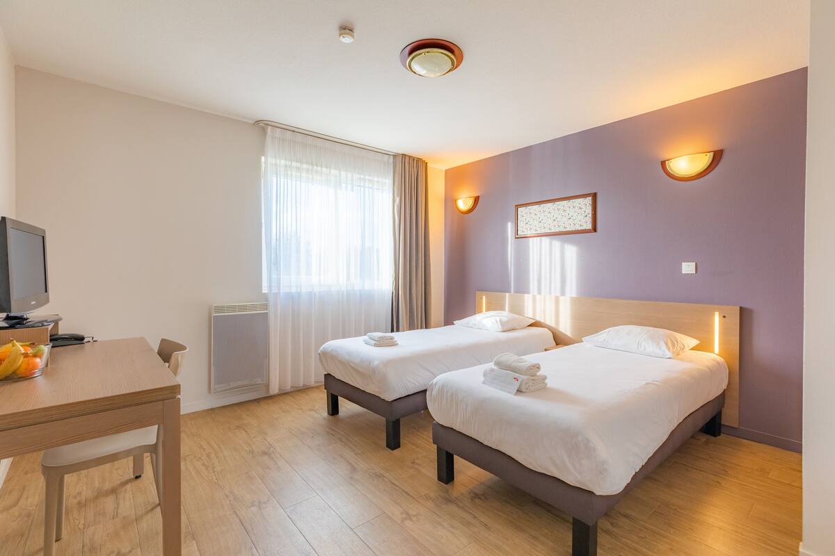 A hotel room with two beds, a desk with a chair, a television, and a window with curtains.