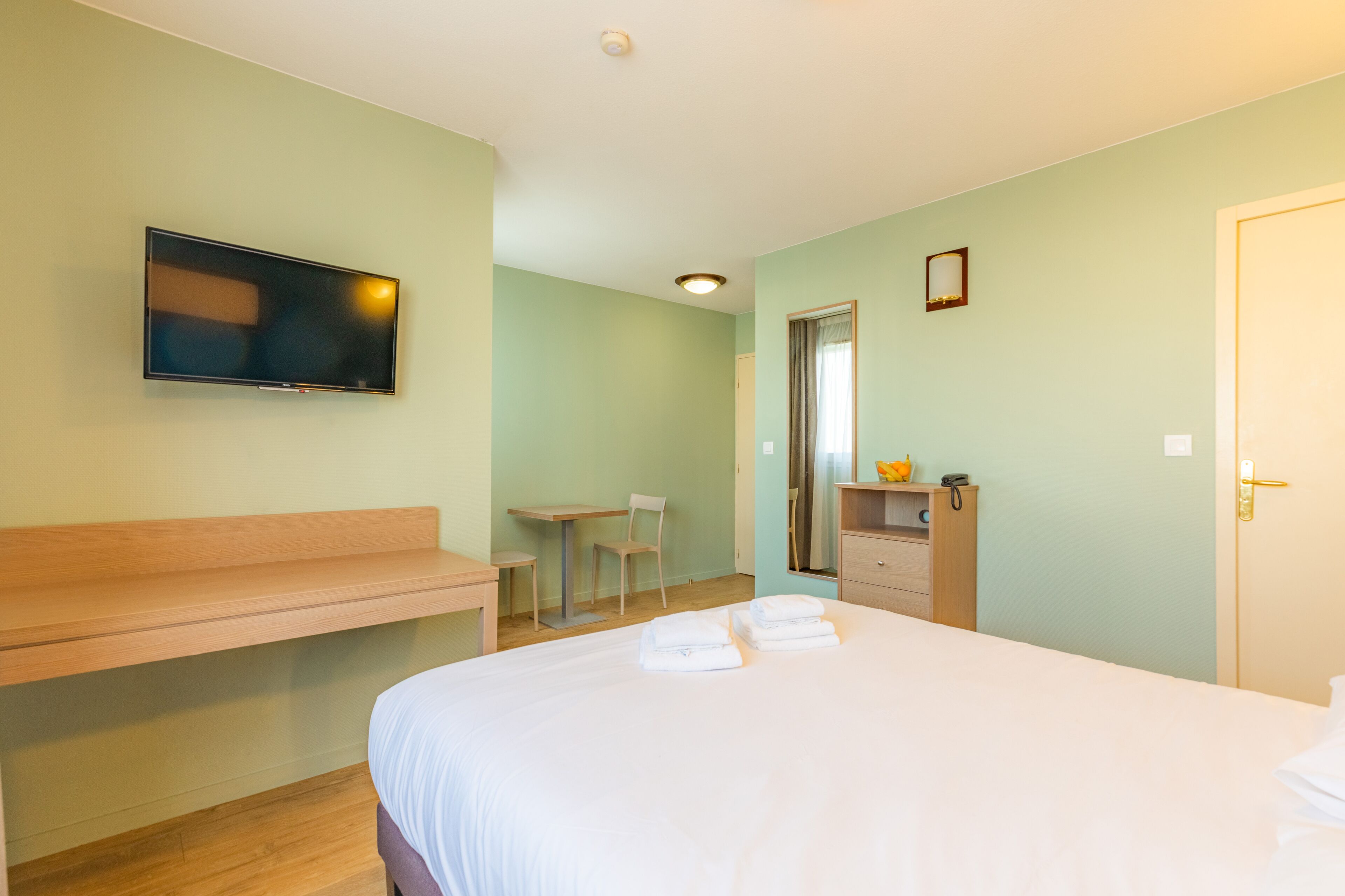 A hotel room with a bed, a desk, a chair, a TV, a mirror, and a door.