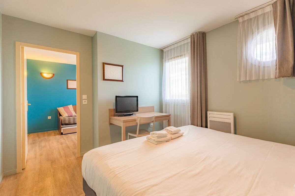 A hotel room with a bed, a desk with a television, a chair, and a small seating area with a couch and pillows.