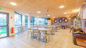 Eine großzügige Cafeteria mit mehreren Tischen und Stühlen, großen Fenstern und einem an der Wand befestigten Feuerlöscher.