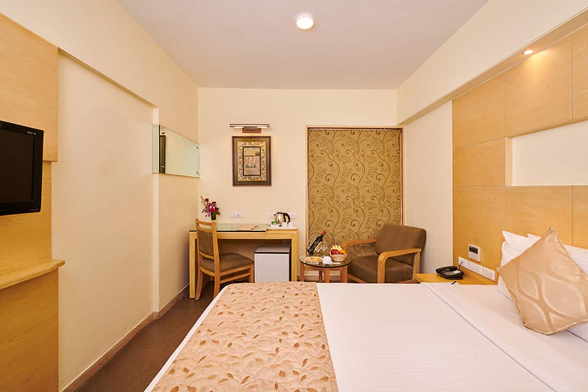 Habitación de hotel con cama, escritorio, silla, televisor y una mesita con un jarrón de flores.