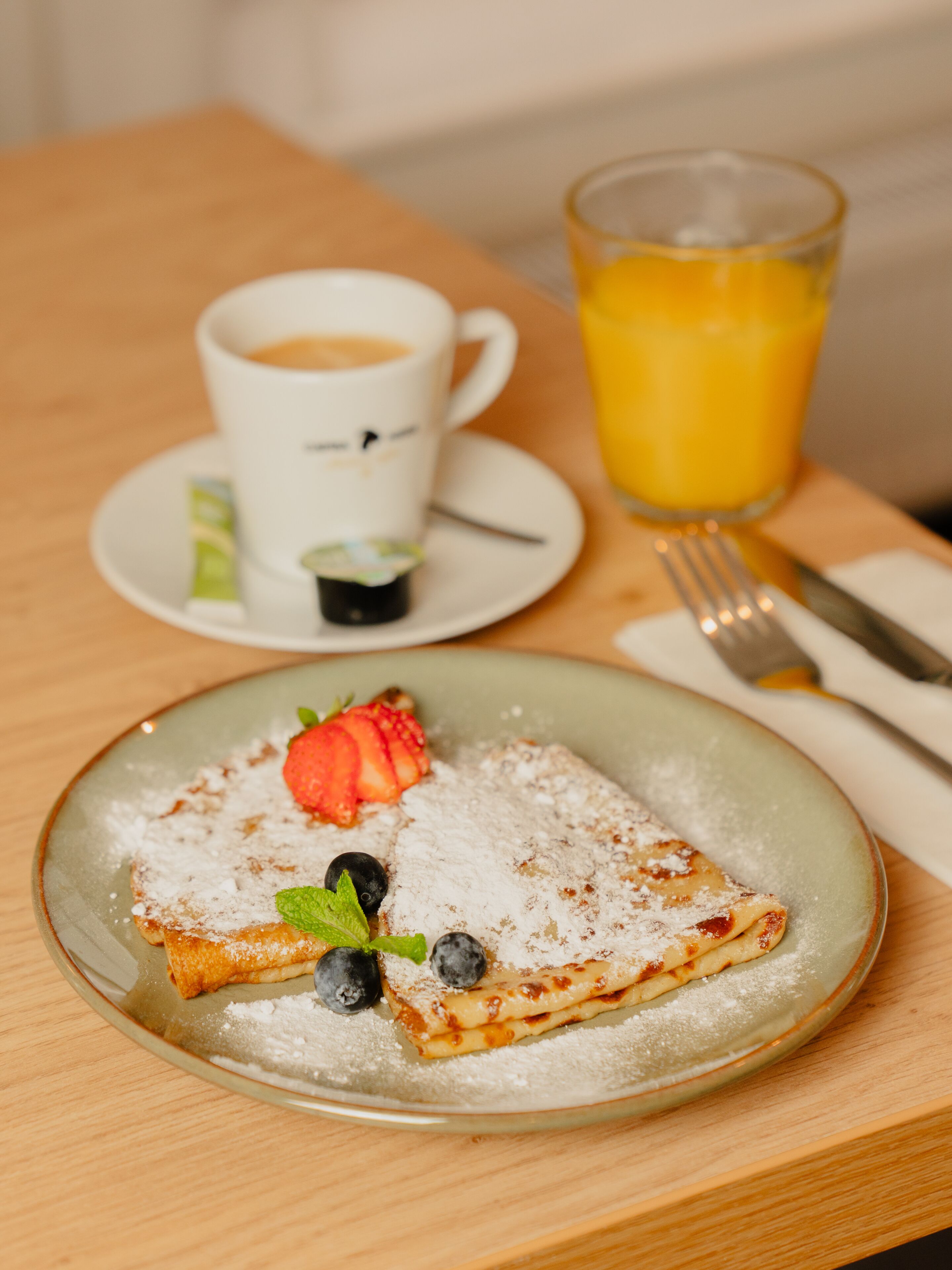 Un desayuno con una fuente de tortitas cubiertas de fresas, arándanos y una hoja de menta, una taza de café y un vaso de zumo de naranja sobre una mesa de madera.