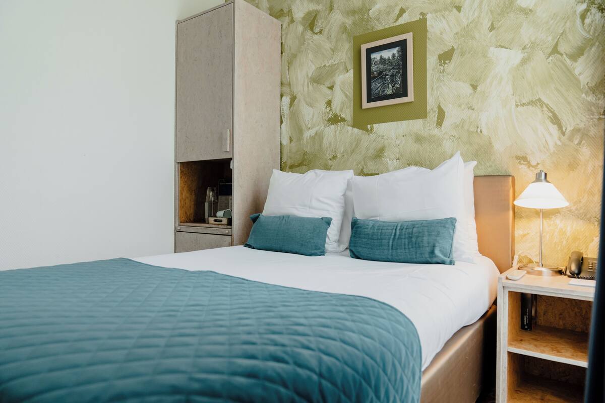 A neatly made bed with a teal quilt, a bedside lamp, and a small nightstand with a phone and a book.