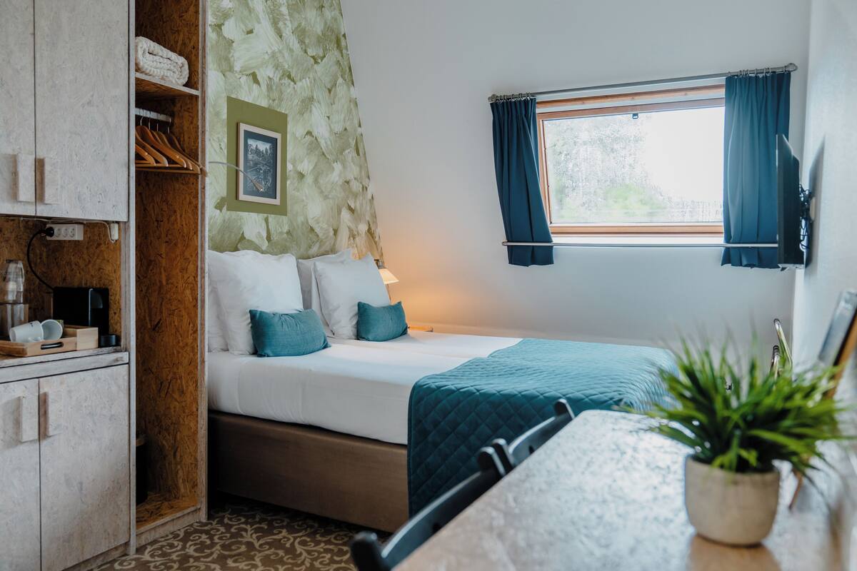 A bedroom with a bed, a wooden wardrobe, a small table, a potted plant, and a window with curtains.