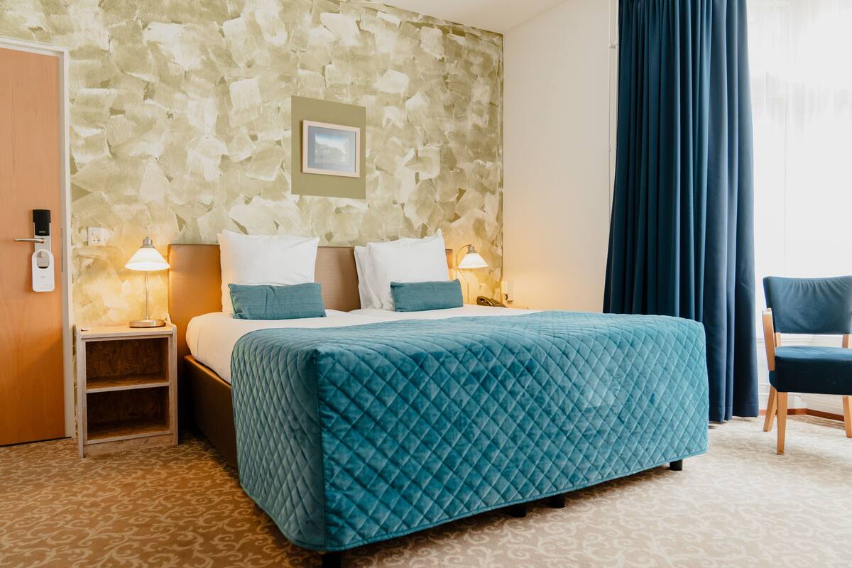 A hotel room with a stone-patterned wall, a bed with a quilted headboard, two bedside tables with lamps, a chair, and a door.