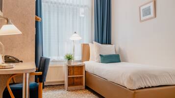 A hotel room with a bed, a desk with a lamp, a chair, a small table with a plant, and a window with curtains.