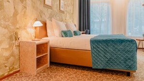 Habitación de hotel con cama, mesita de noche con lámpara, una silla y un gran ventanal con cortinas.