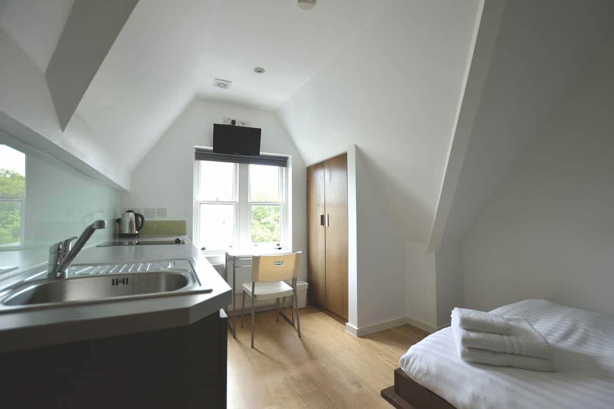 A bedroom with a bed, a window, a desk, and a kitchenette.
