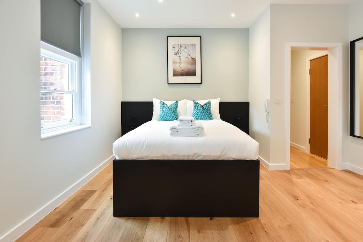 A modern bedroom with a large bed, a window, a framed picture on the wall, and a wooden floor.