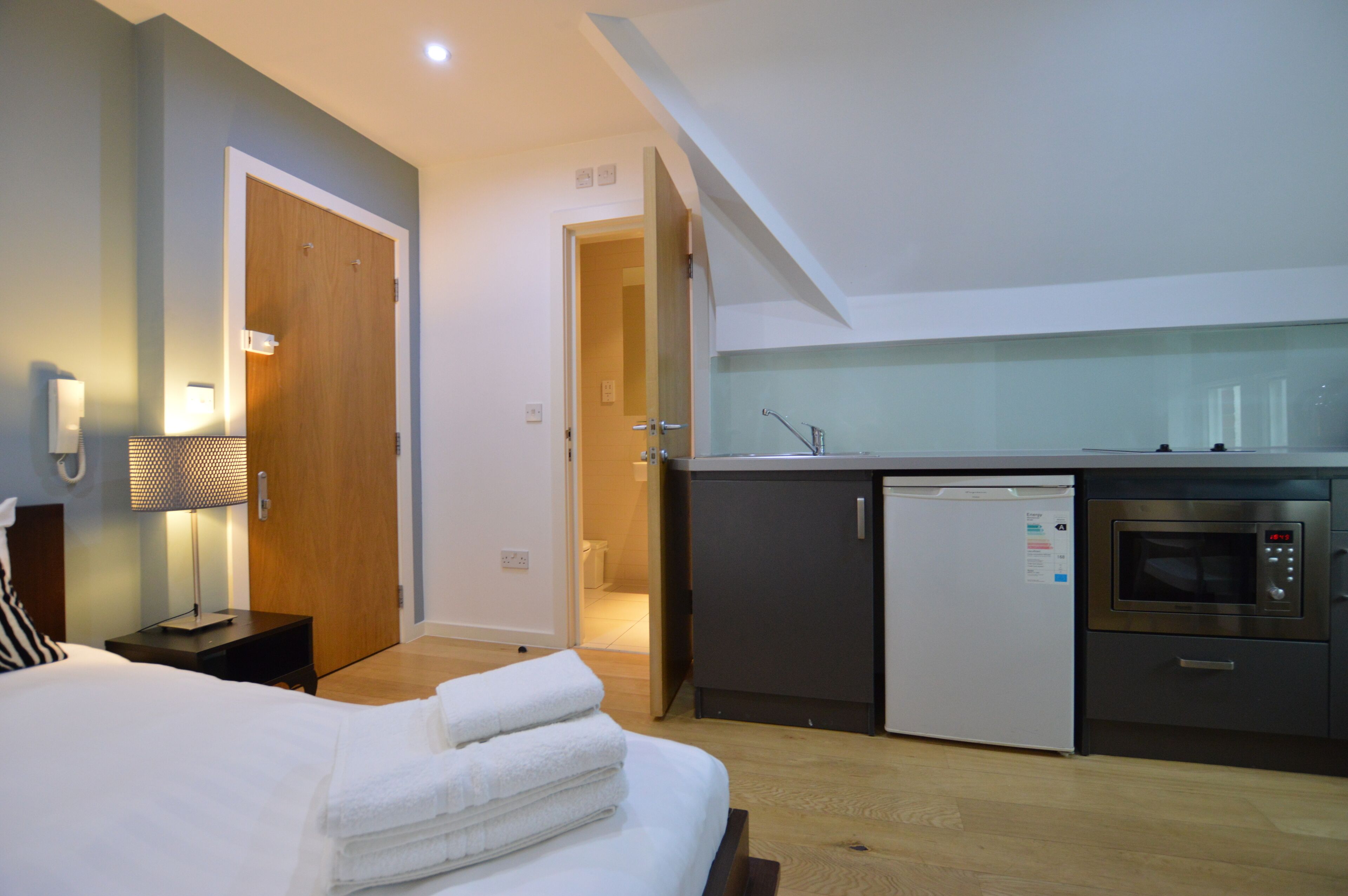 A compact hotel room with a bed, bedside lamp, and a kitchenette area including a refrigerator and oven.