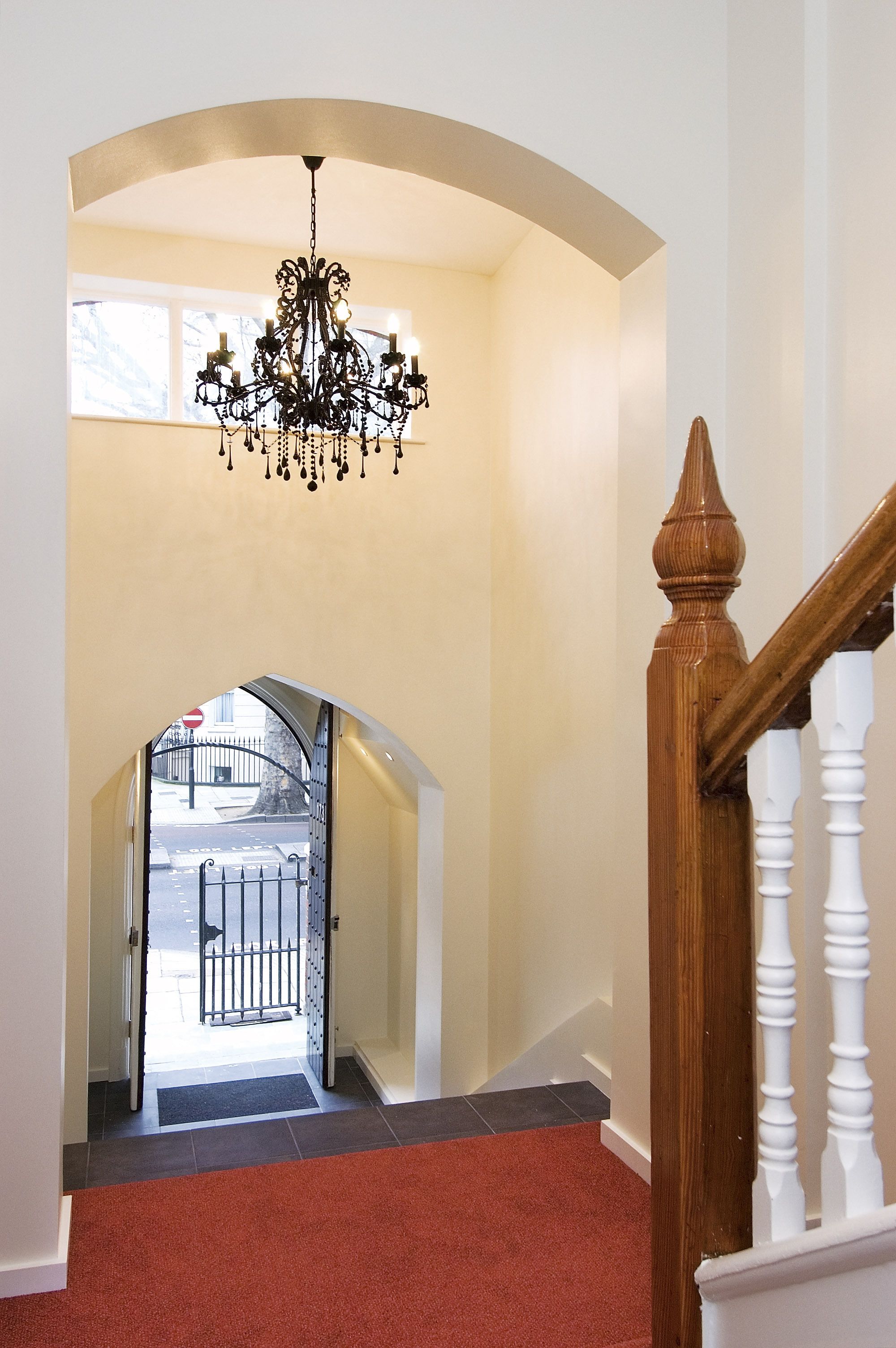 A hallway with a chandelier, a wooden staircase, and a view of the street through an archway.