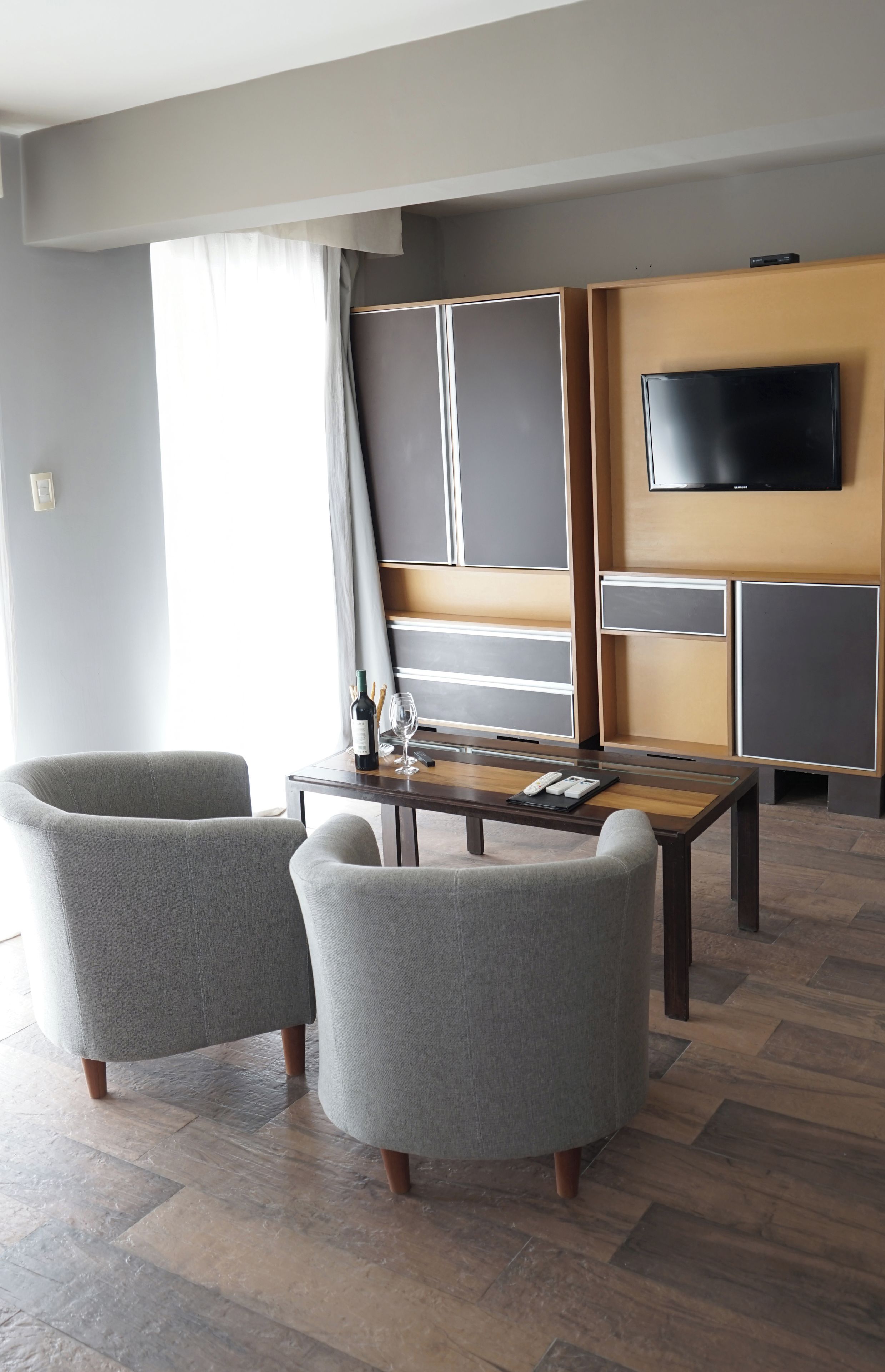 Una habitación de hotel moderna con un televisor de pantalla plana montado en la pared, una mesa de centro de madera, dos sillones grises y un ventanal con cortinas.