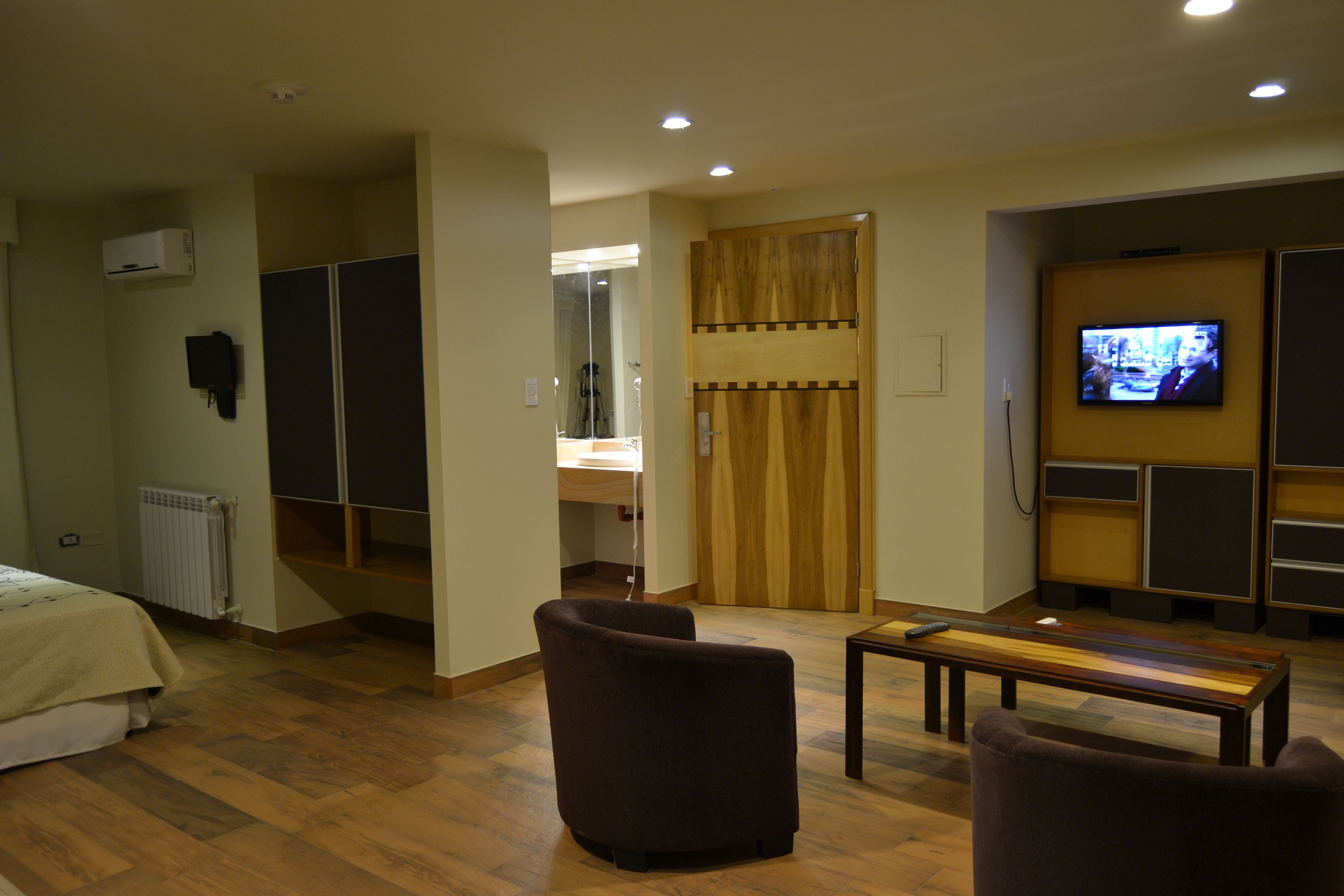 Een hotelkamer met een bed, twee fauteuils, een houten tafel, een televisietoestel, een kledingkast en een spiegel.