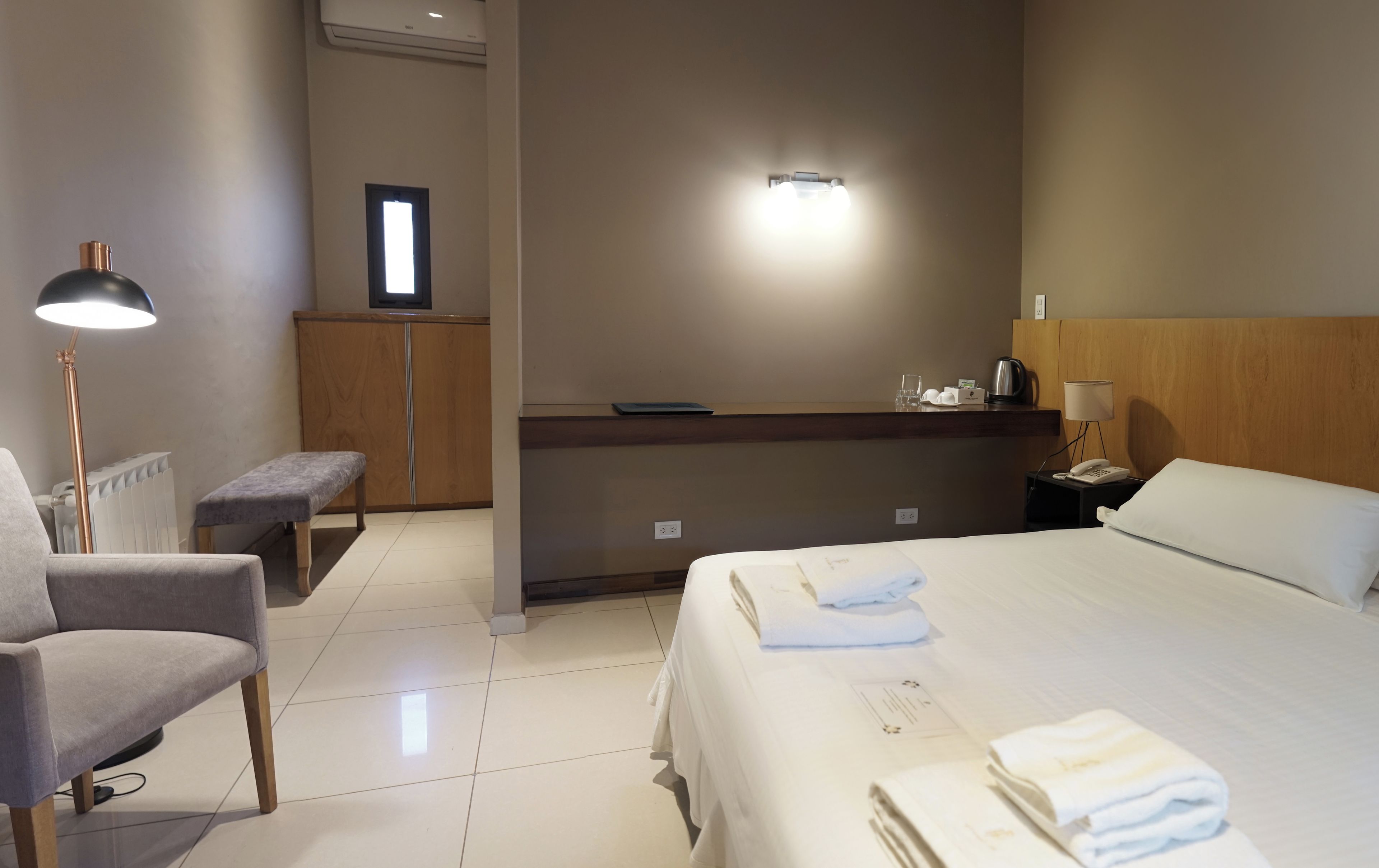 Una habitación de hotel con cama, escritorio, silla, lámpara y radiador.