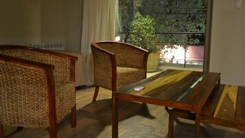 Een kamer met een houten tafel en twee rieten stoelen, een raam met uitzicht op bomen, en een balkon met een hekwerk.
