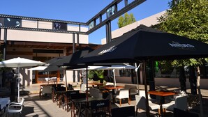 An outdoor café with tables and chairs under umbrellas, a building with large windows, and a tree in the background. - Plaza Paradiso by Camberland (Chacras de Coria)