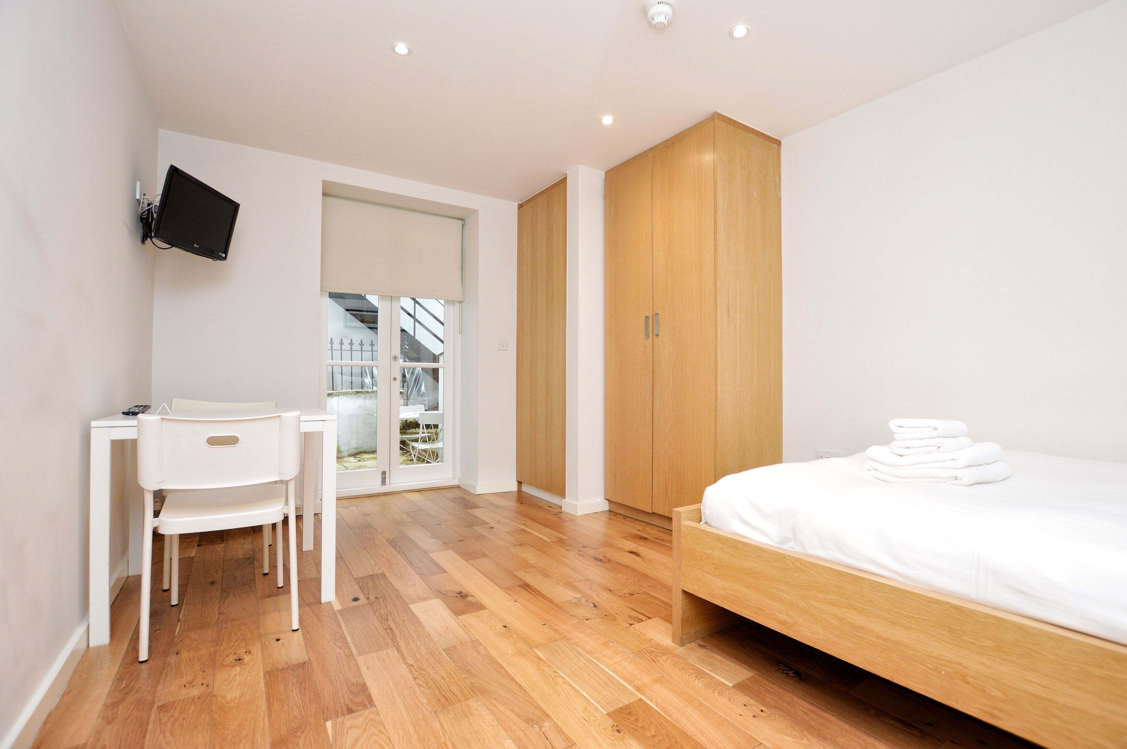 A bedroom with a wooden bed, a desk with a chair, a TV mounted on the wall, and a wardrobe.