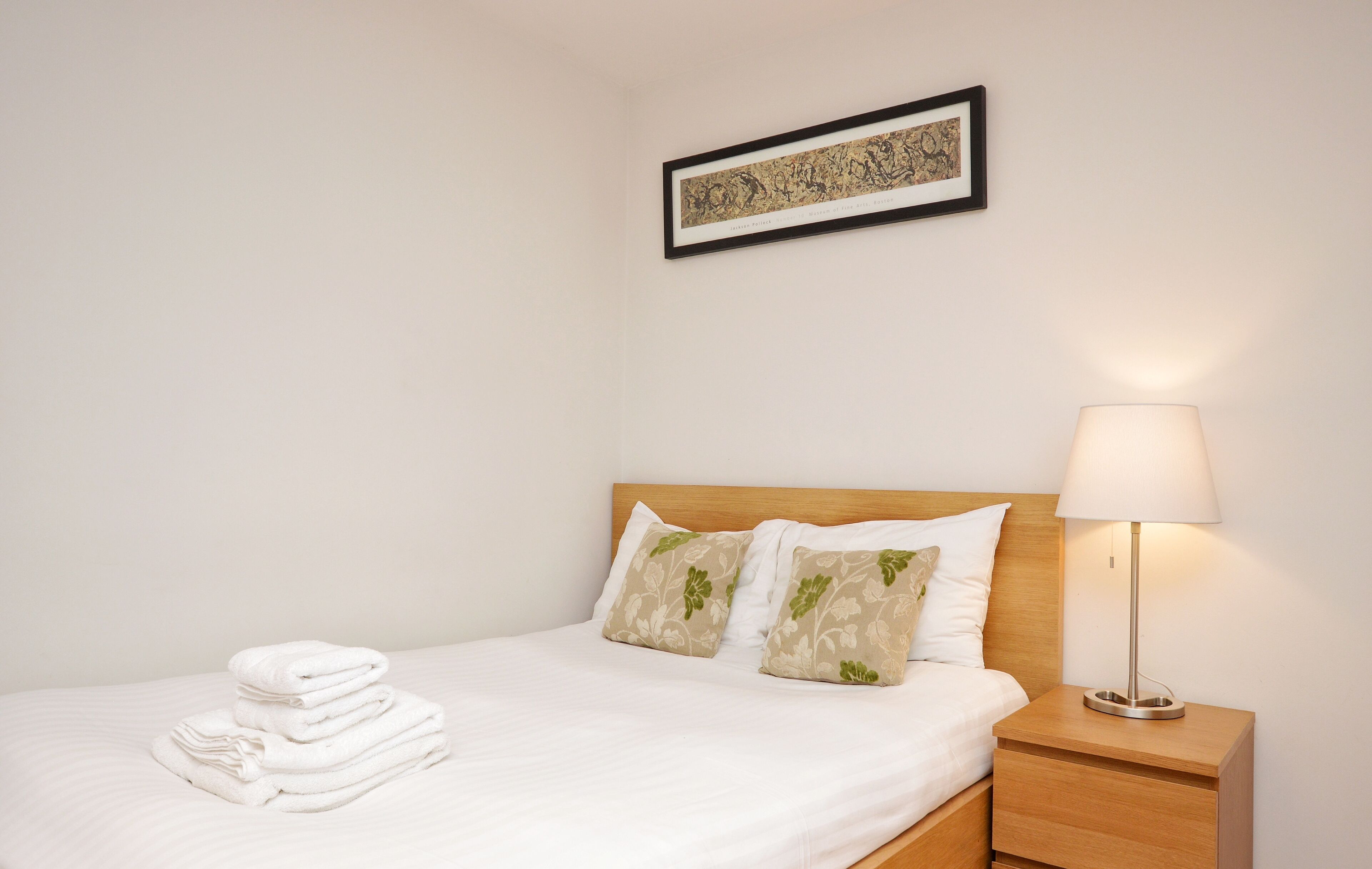 A neatly made bed with white linens, two decorative pillows, a folded towel, a bedside lamp, and a framed artwork on the wall.