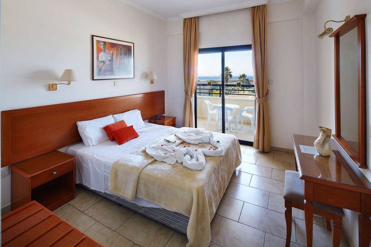Una habitación de hotel con una cama, una mesita de noche de madera, un escritorio, una silla, un ventanal con vistas a la playa y un cuadro en la pared.