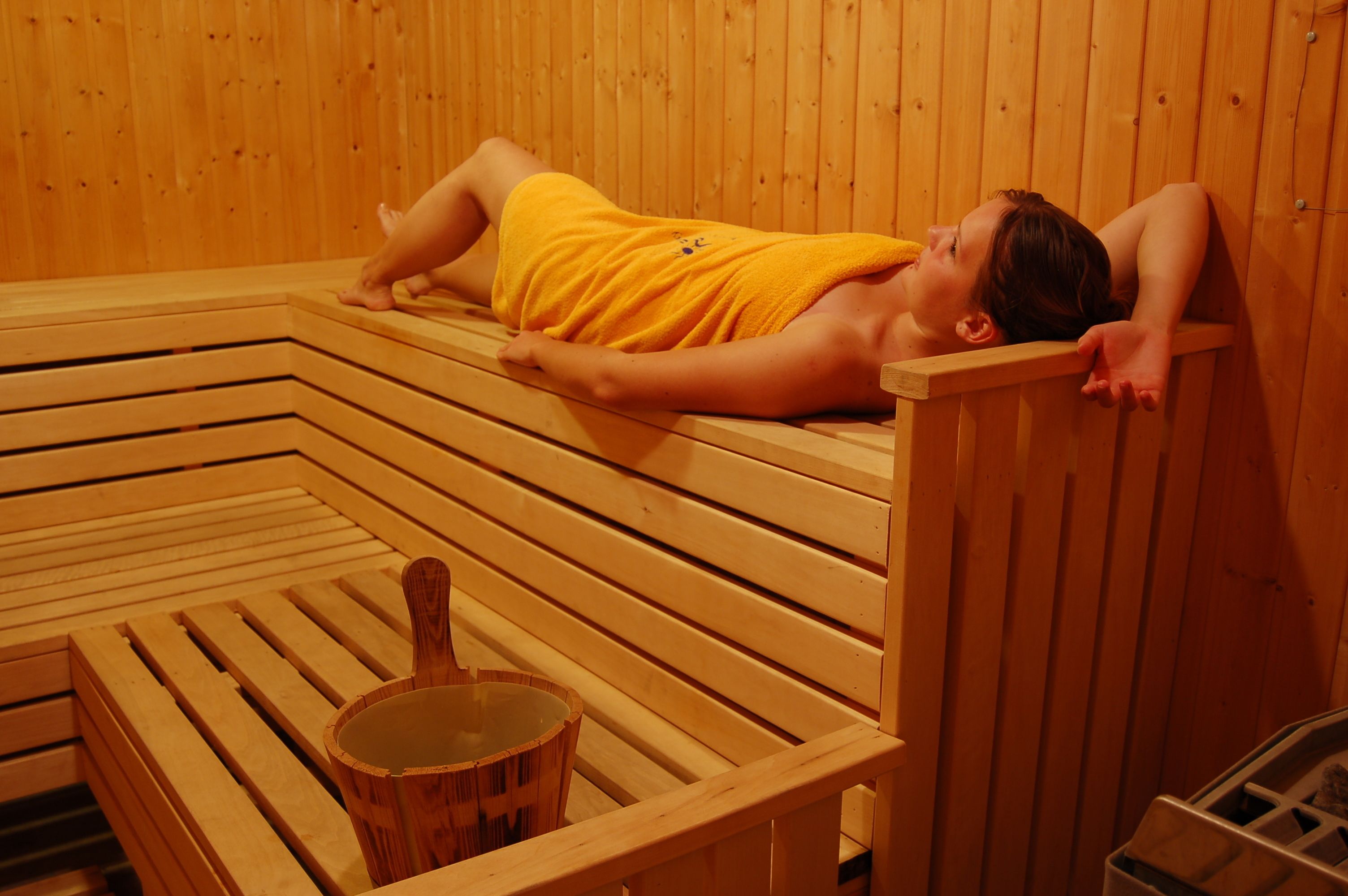 A person lying on a wooden bench in a sauna room with a wooden bucket.