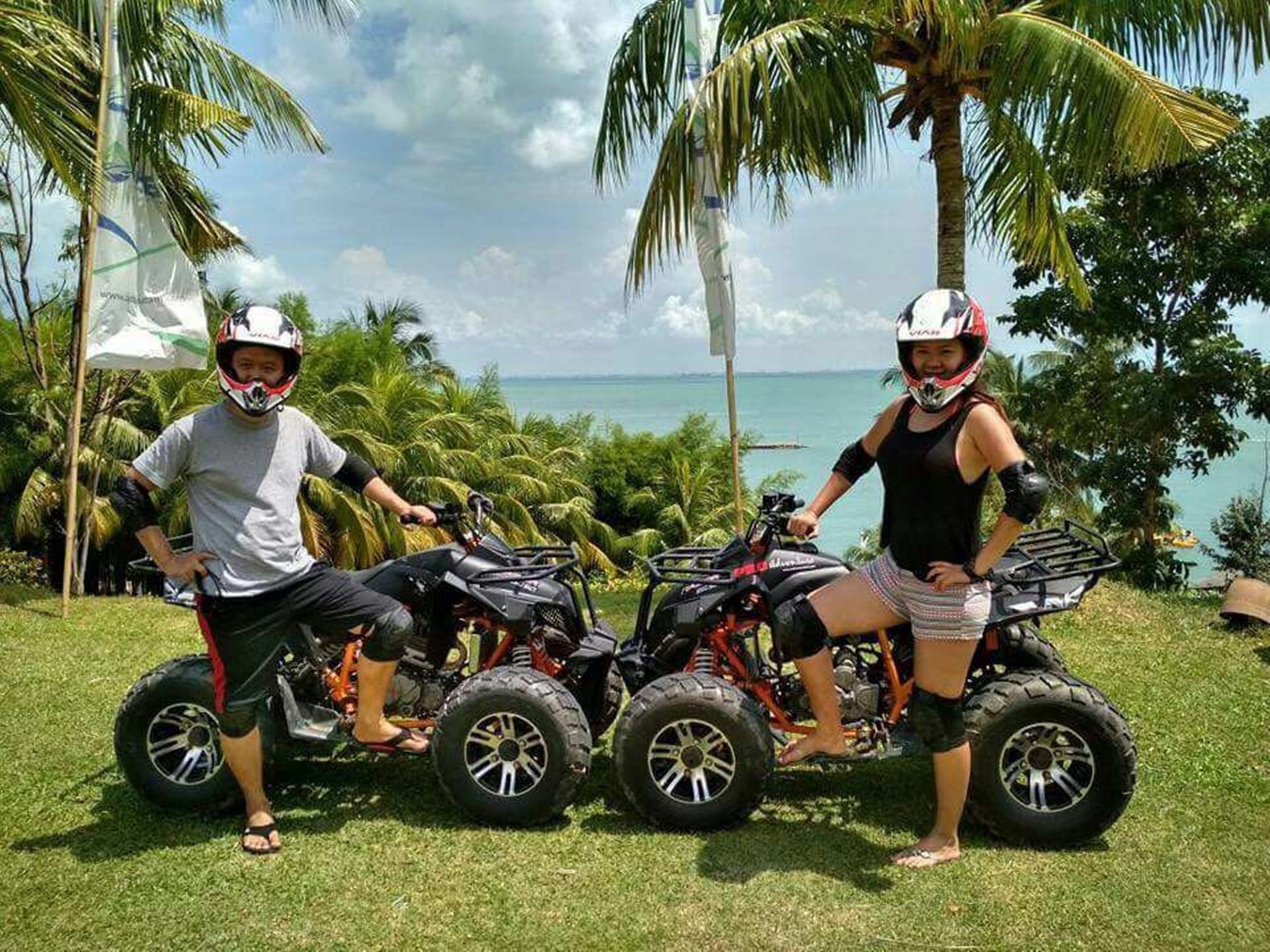 Two people on quad bikes in a tropical setting with palm trees and a view of the ocean.