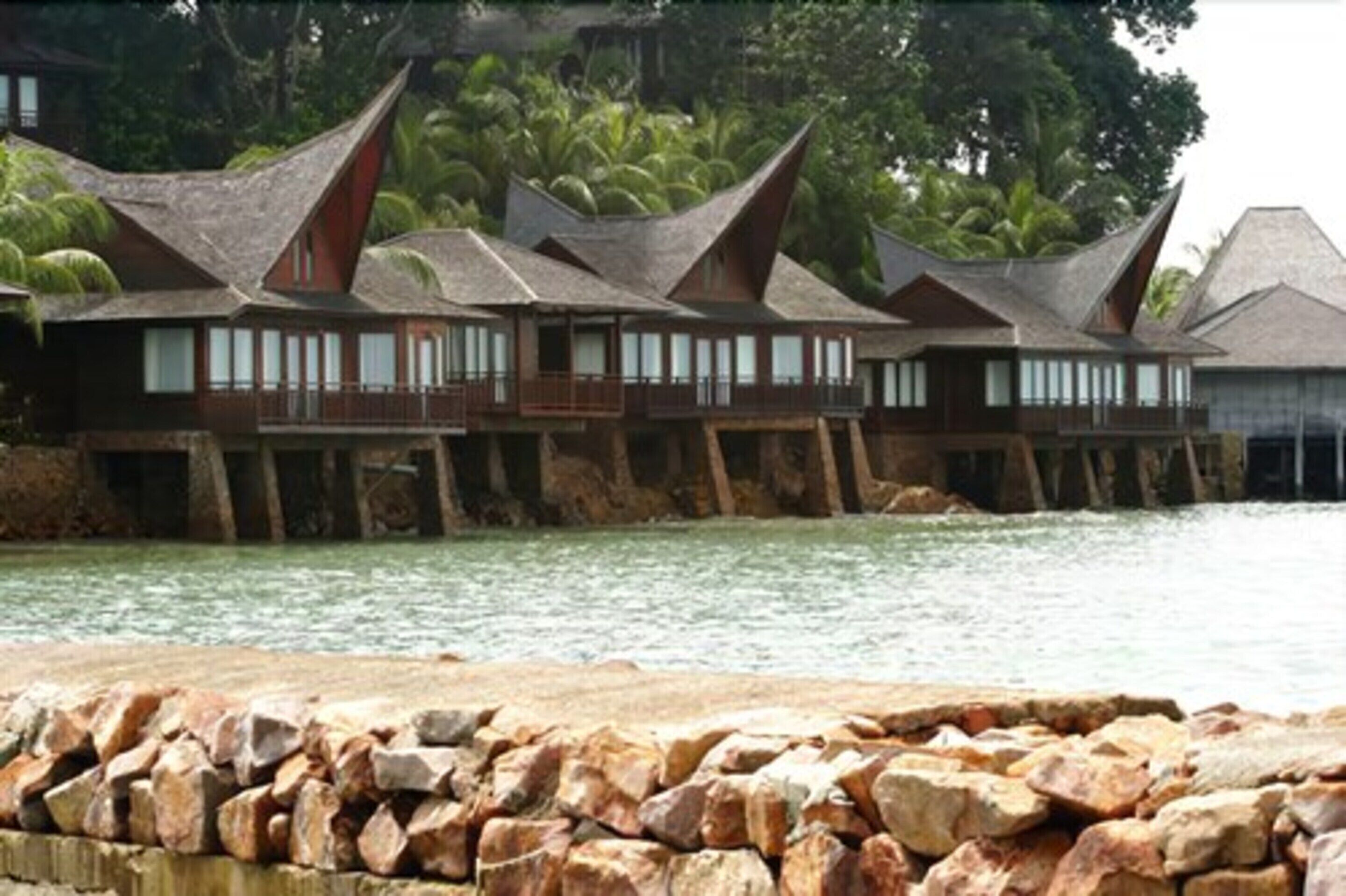A row of wooden overwater bungalows with balconies, supported by stilts over water, surrounded by lush greenery.
