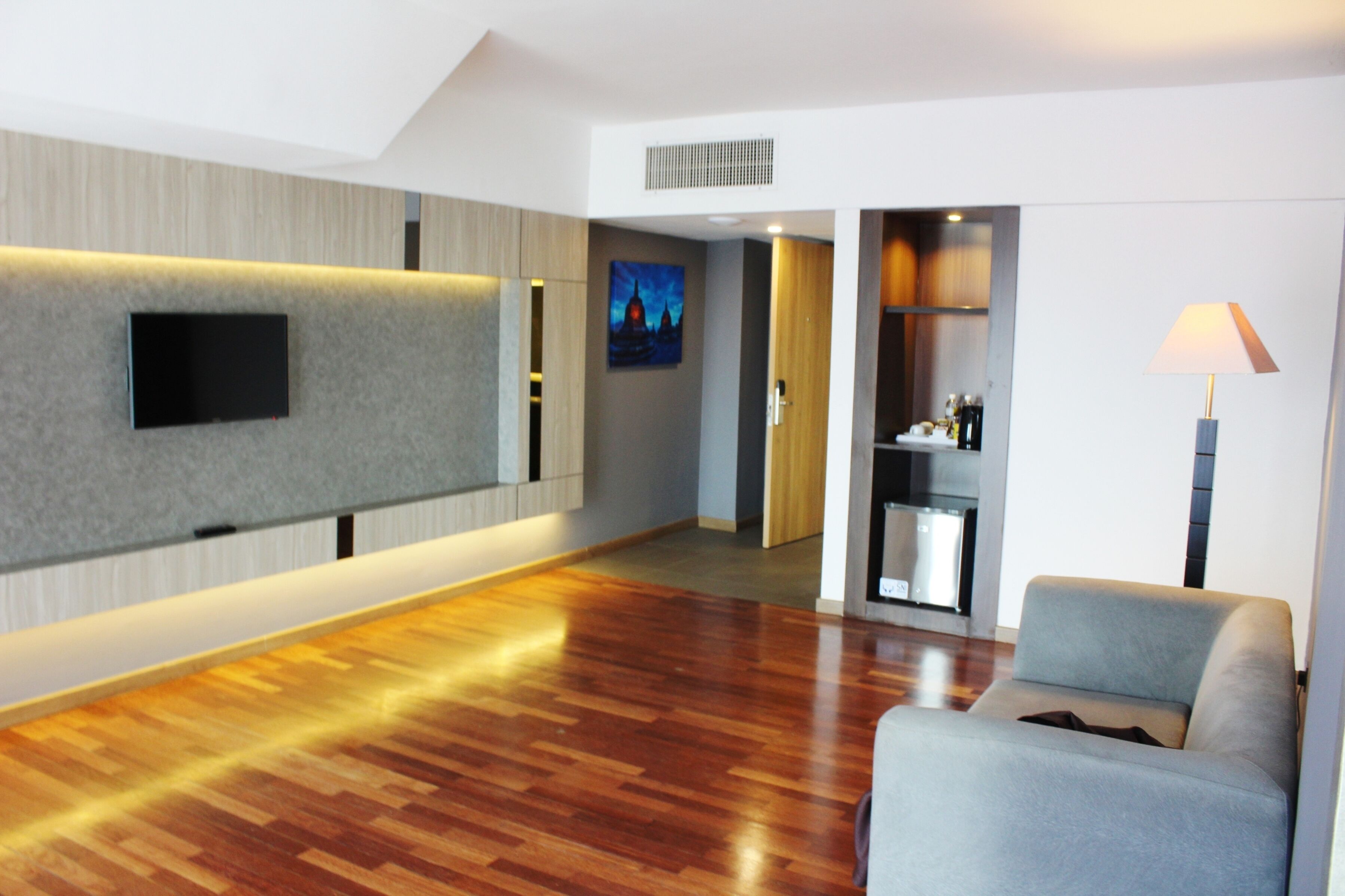 A modern hotel room with a flat-screen TV mounted on the wall, a wooden floor, a grey sofa, and a lamp.