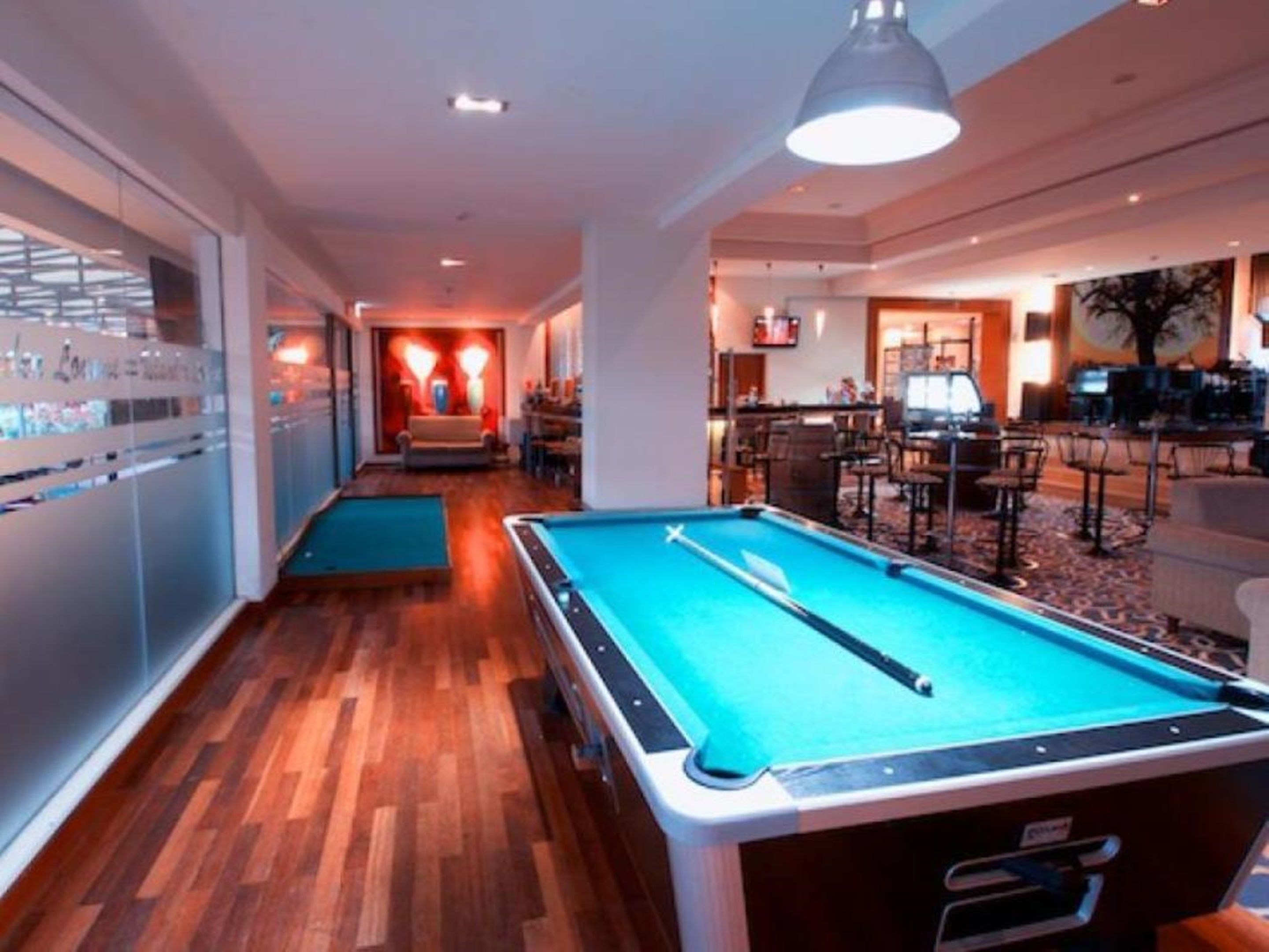 A pool table in a hotel lobby with a wooden floor, a bar area, and a seating area with chairs and tables.