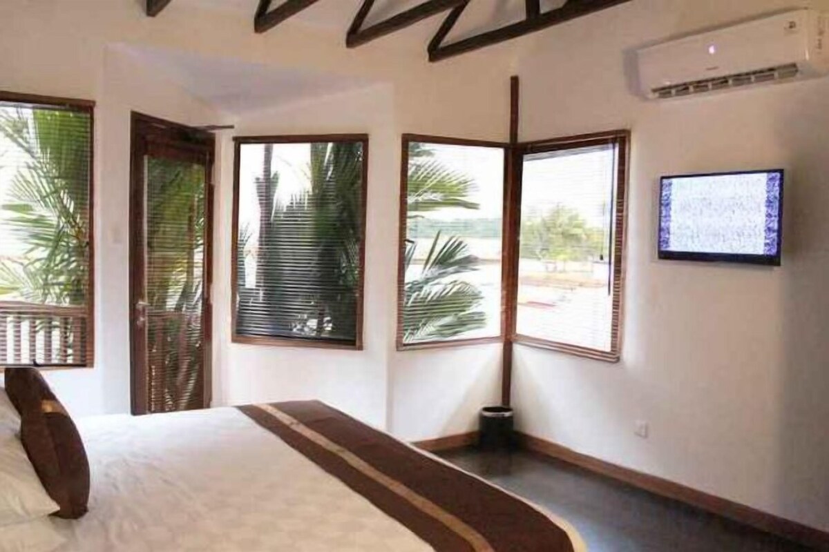 A hotel room with a bed, large windows, a television, and a view of palm trees.
