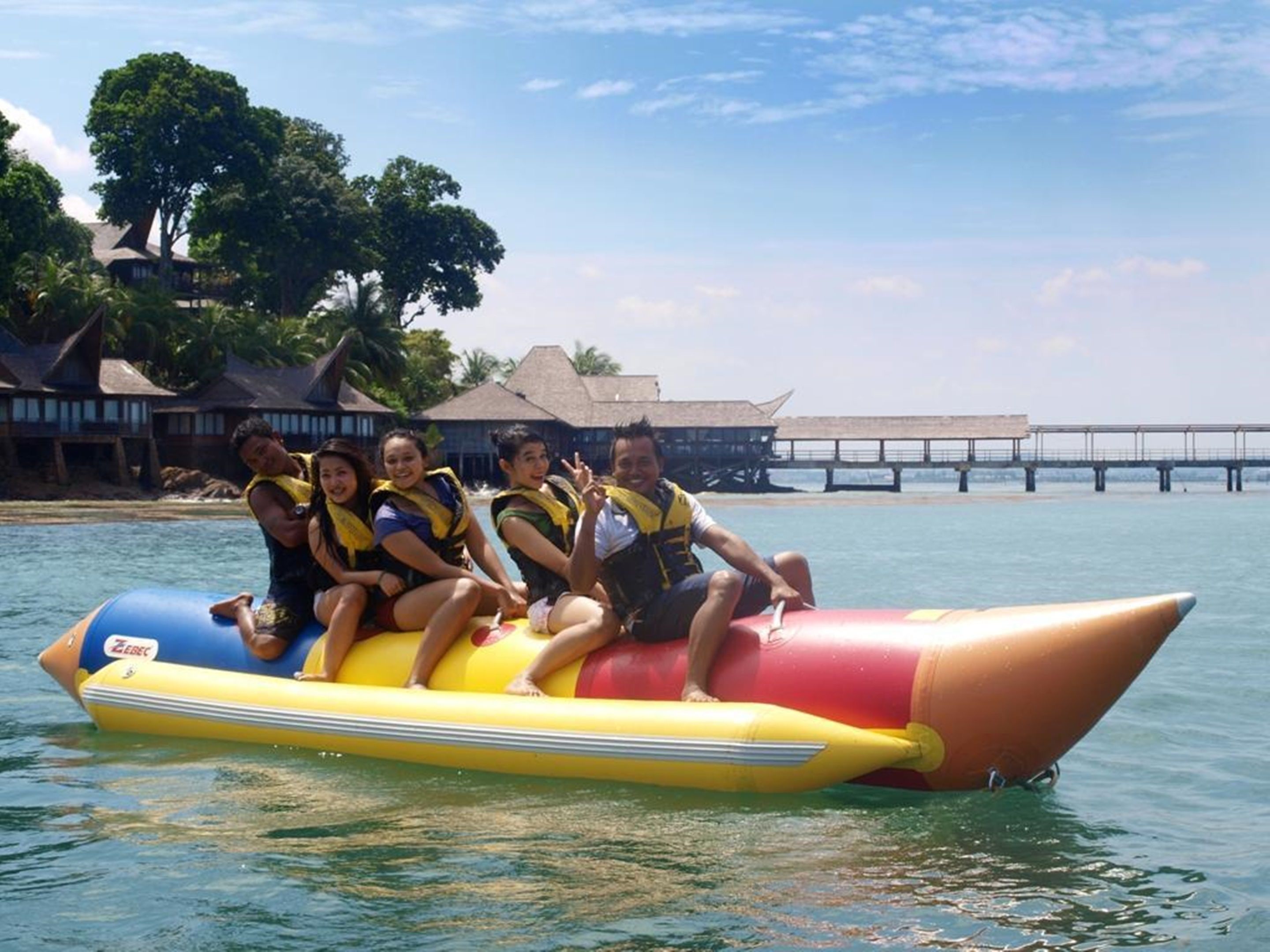 A group of people on a yellow and red inflatable raft in a body of water with a pier and overwater bungalows in the background.