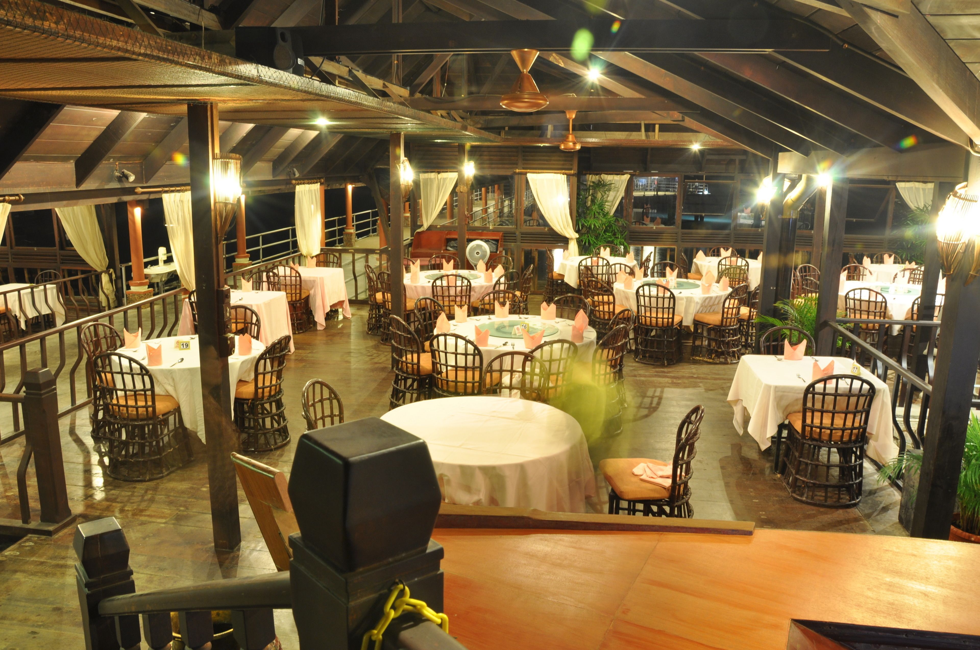 A restaurant with round tables and wicker chairs, set in a spacious indoor area with a high ceiling and exposed beams.