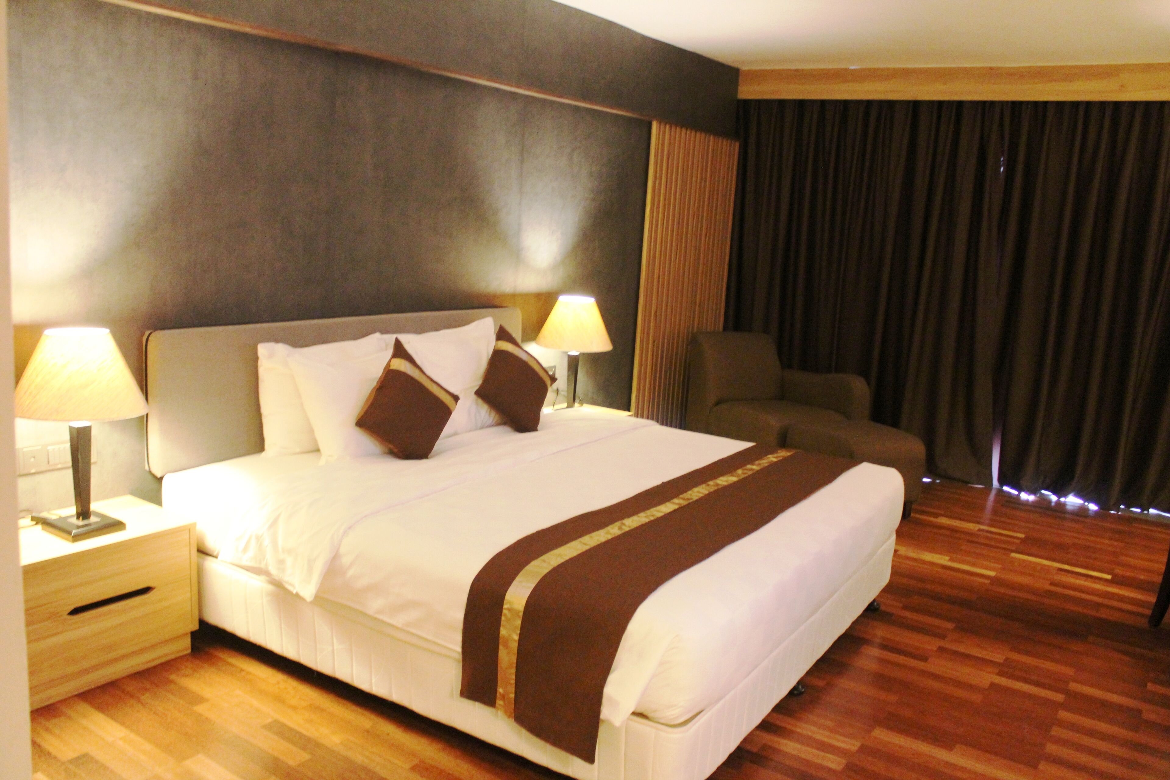A hotel room with a large bed, two bedside tables with lamps, a chair, and a view of the outside through a window with curtains.