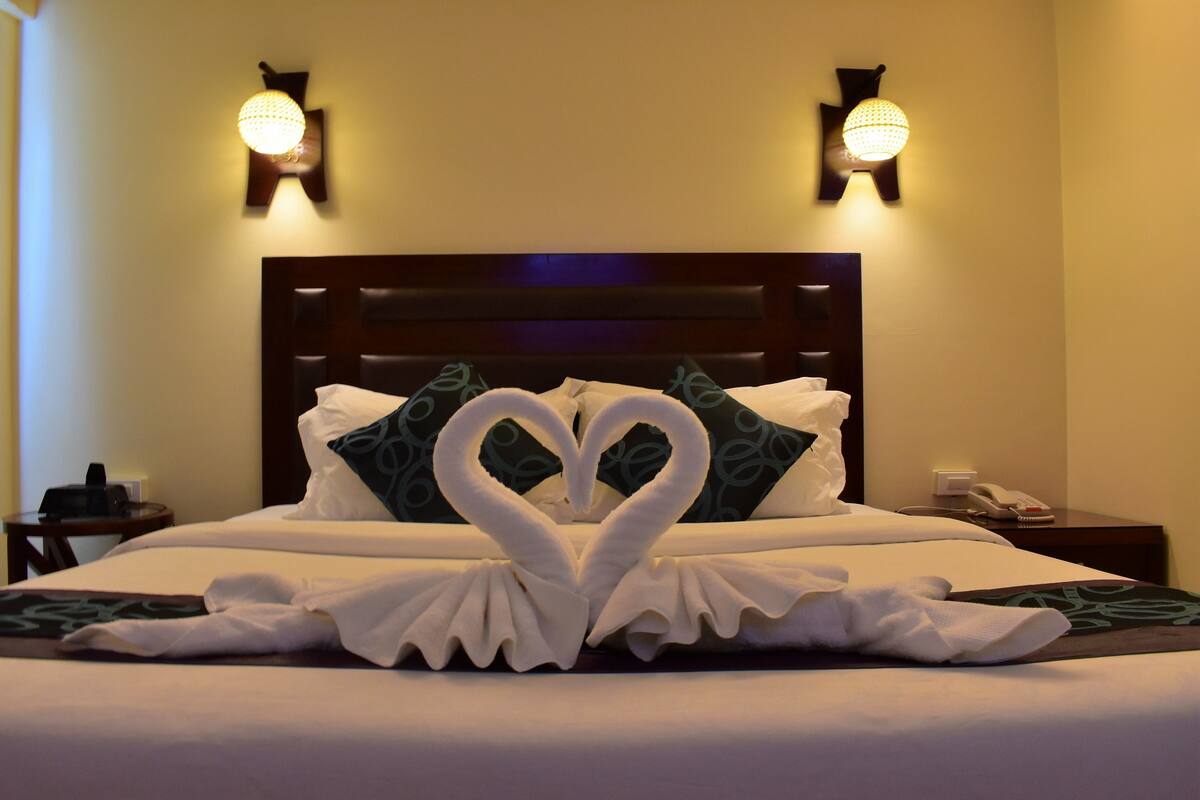 Een hotelkamer met een bed, versierd met handdoeken in de vorm van een hart, een houten hoofdeinde en twee wandlampen.