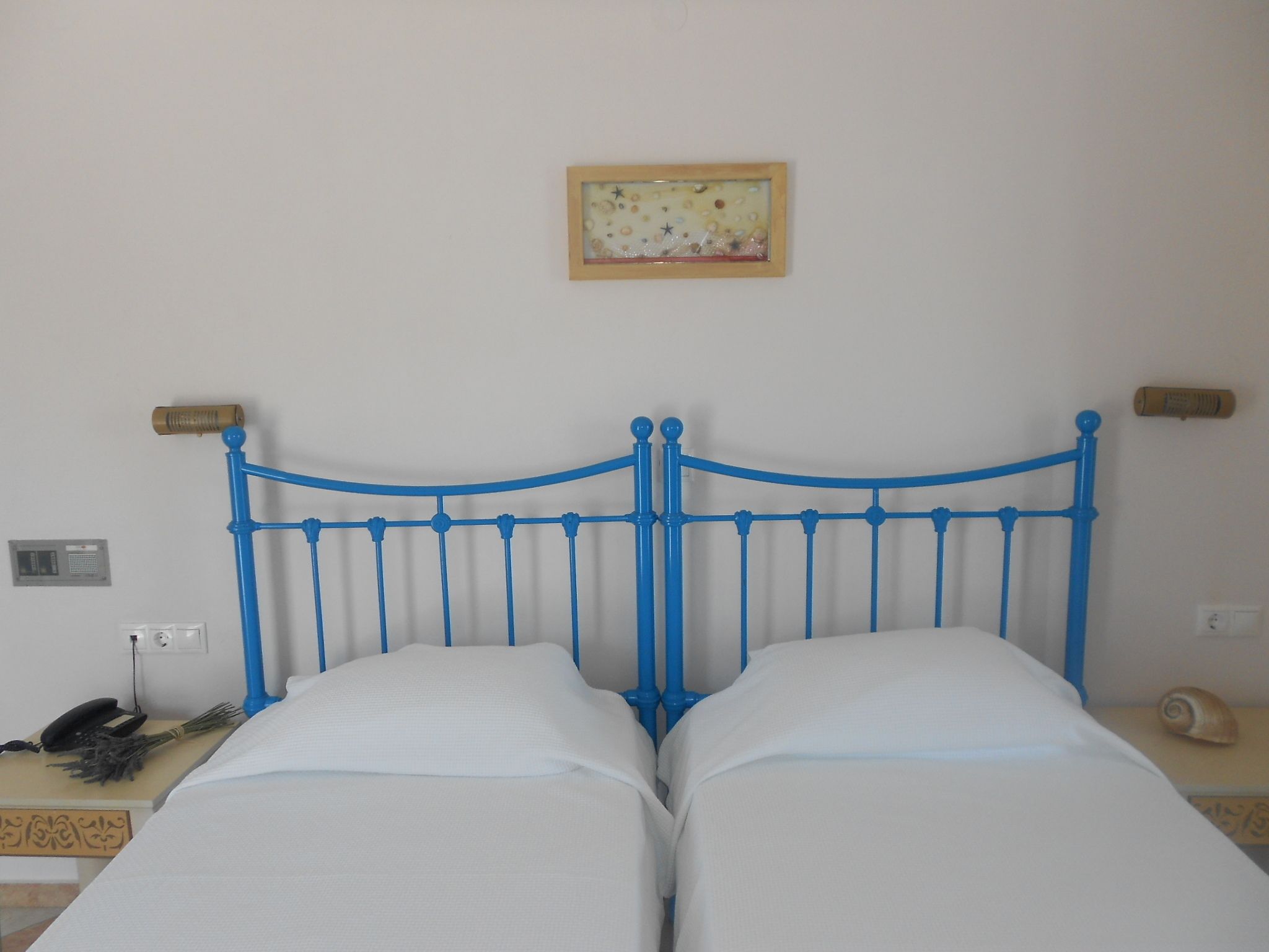 Ein Bett mit blauem Metallgestell und weißer Bettwäsche in einem Zimmer mit schlichter Wand, einem kleinen Bild und einem Nachttisch mit Lampe und Telefon.