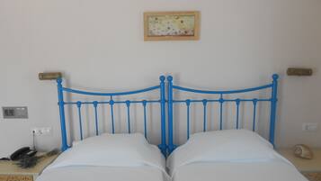 Een bed met een blauw metalen frame en witte beddengoed in een kamer met een effen muur, een klein schilderij en een nachtkastje met een lamp en een telefoon.