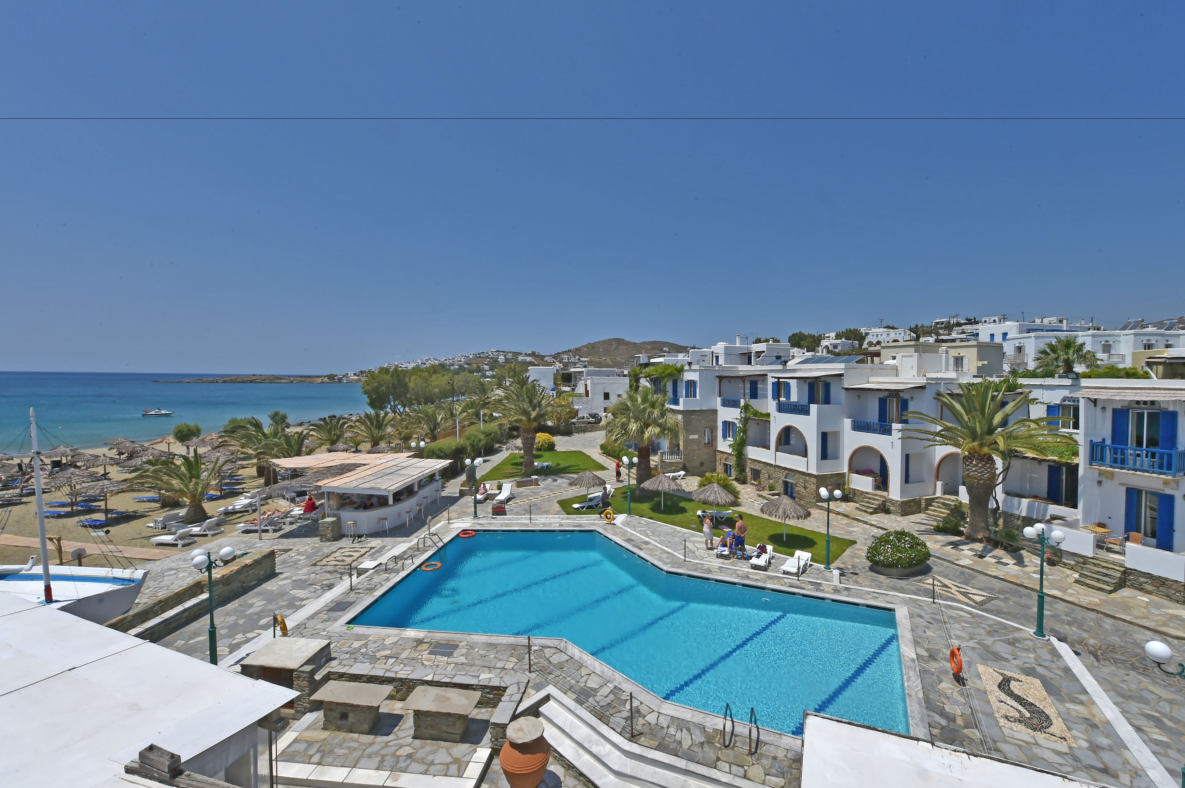 Zona de piscina con vista despejada al mar, rodeada de edificios blancos con detalles azules, y una pequeña playa con sombrillas.